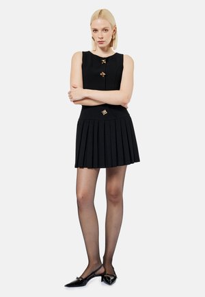 Blondynka stoi ze skrzyżowanymi rękami, ubrana w czarną sukienkę bez rękawów z złotymi guzikami, plisowaną spódnicę, przezroczyste czarne rajstopy i czarne sandały na obcasie typu slingback.