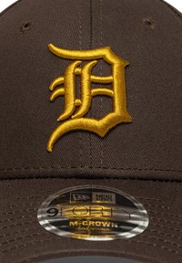 Braune Baseballmütze mit einem großen, goldenen, gestickten "D"-Logo auf der Vorderseite und einem New Era 9FORTY M-CROWN-Aufkleber auf dem Schirm.
