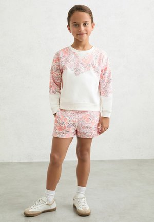 Jauna meitene stāv ar rokām šortos, valkājot baltu džemperi ar rozā ziedu dizainu, atbilstošus ziedu šortus un baltas sporta kurpes.