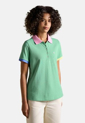 Žena s kučeravými vlasmi, ktorá nosí zelenú polokošeľu s ružovým golierom a farebnými manžetami na rukávoch, v kombinácii s svetlými nohavicami.