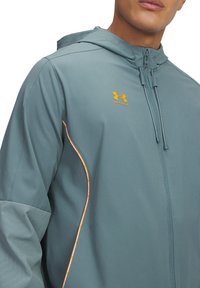 Letvægts, teal regnjakke med hætte. Har orange detaljer og et Under Armour-logo på brystet. Glat tekstur, lynlås lukning.