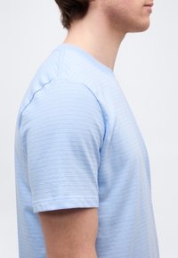 Πλευρική όψη ατόμου που φοράει ανοιχτό μπλε πλεκτό πουκάμισο με κοντά μανίκια, με υφή, σε απλό λευκό φόντο.