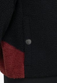 Veste en polaire avec un corps noir et des accents rouges, matériau texturé et détail de logo en métal. Conçue avec une coupe décontractée et des poignets élastiques.