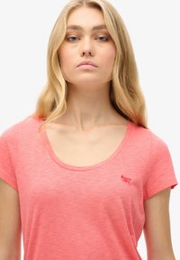 Ung kvinde med langt blondt hår iført en koral pink T-shirt med rund hals og en lille rød "Superdry"-logo på brystet.
