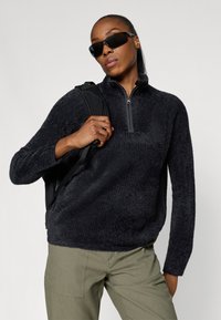 Sort fleece trøje med halv-zip krave, struktureret overflade og lange ærmer, parret med olivengrønne bukser og en sort rygsæk.