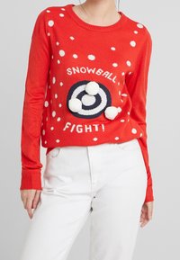 Pull rouge à pois blancs, avec un motif circulaire bleu marine et des pompons blancs. Poignets côtelés et encolure ronde.