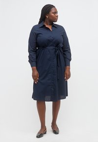 Γυναίκα που στέκεται, φορώντας ένα μπλε σκούρο φόρεμα-πουκάμισο με κουμπιά και δεμένη ζώνη στη μέση, καθώς και μαύρα παπούτσια με χαμηλό τακούνι, κοιτάζει προς τα δεξιά της.