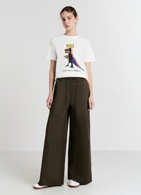 T-shirt blanc à manches courtes avec un graphique coloré de dinosaure et du texte ; associé à un pantalon large vert olive et des baskets blanches.