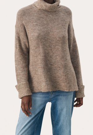 Pull à col roulé marron en tricot avec poignets repliés porté avec un jean bleu délavé sur une personne devant un fond uni.