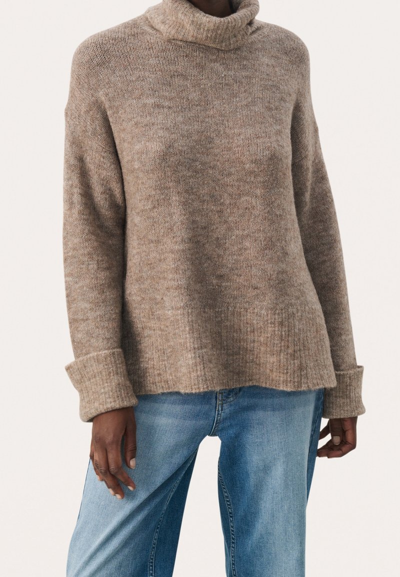 Pull à col roulé marron en tricot avec poignets repliés porté avec un jean bleu délavé sur une personne devant un fond uni.
