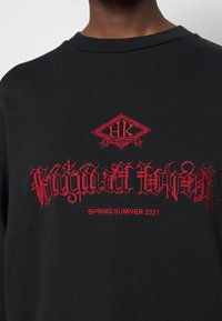 Svart sweatshirt med röd broderad text. Designen inkluderar en diamantformad logotyp med intrikata detaljer och "SPRING SUMMER 2021" under.