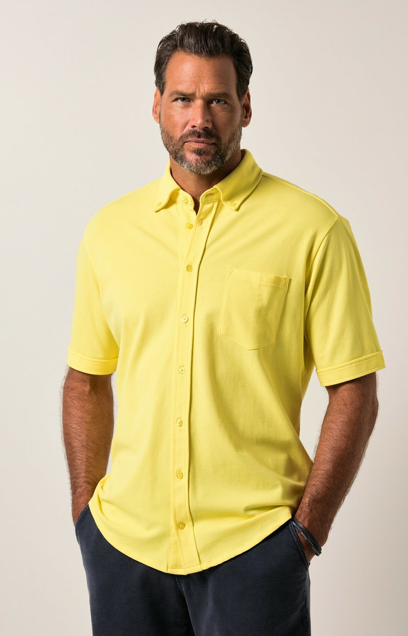 Camiseta polo amarilla de manga corta con botones al frente, cuello y un bolsillo en el pecho. Textura suave y corte relajado, combinada con pantalones oscuros.