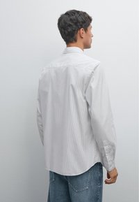 Wit gestreept overhemd met lange mouwen, een gebogen zoom en knoopmanchetten, gecombineerd met lichtblauwe denim jeans, gezien van achteren.