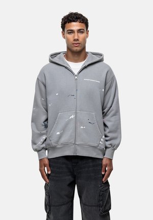 Sweat à capuche gris zippé avec une poche kangourou, arborant des motifs de gouttes de peinture en bleu et rose, et le texte du logo sur le côté gauche de la poitrine. Coupe régulière.
