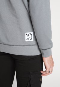 Sudadera gris con puños acanalados, presentando una etiqueta blanca con texto y logotipo en negro, combinada con pantalones negros que destacan detalles en los bolsillos.