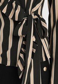 Blouse rayée noire et beige avec un détail volanté et une taille nouée. Le matériau est texturé, avec une finition lisse et un accent en métal visible.