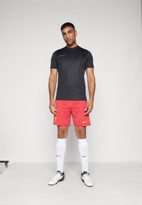 Must musta särk musta kuldse Nike logo ja punased lühikesed püksid Liverpooli embleemiga, valged sokid ja mustad ja valged jalatsid.
