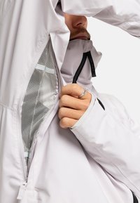 Mann trägt eine hellgraue Jacke und zieht ein seitliches Netzbelüftungsfeld auf, wobei ein Teil seines Gesichts und seiner Hand mit einem gemusterten Ring sichtbar ist.