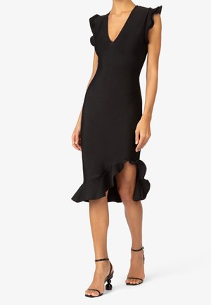 Mujer que lleva un vestido negro sin mangas con bajo asimétrico con volantes y sandalias negras de tacón alto con tiras en el tobillo.