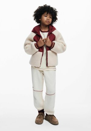 Fleecejacke in Creme und Burgunderrot, mit Reißverschluss und gerippten Bündchen; kombiniert mit hellbeigen Hosen mit kontrastierenden Nähten und braunen Schuhen.