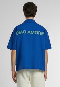 Kortærmet skjorte i livlig blå bomuld; har "CIAO AMORE" i lysegrønne bogstaver på ryggen; afslappet pasform og spidse krave.