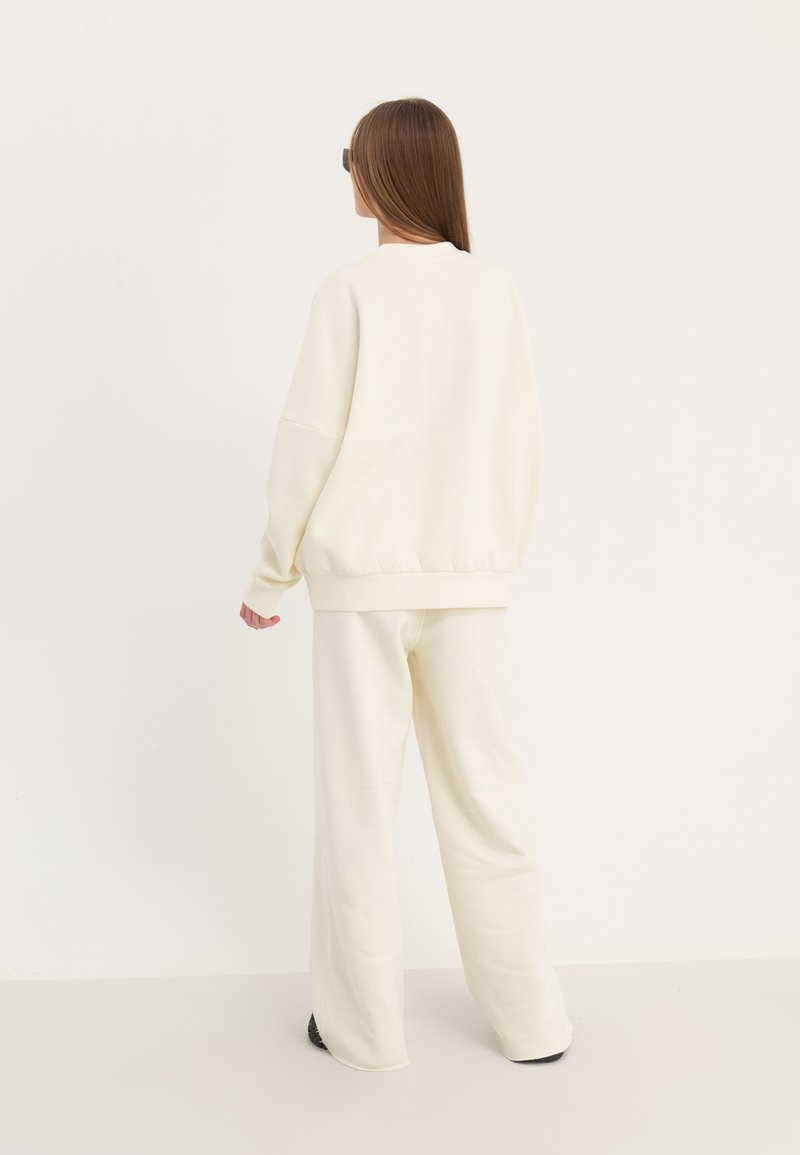 Felpa oversize di colore crema con polsini e orlo a coste, abbinata a pantaloni larghi dello stesso colore, mostrata da dietro.