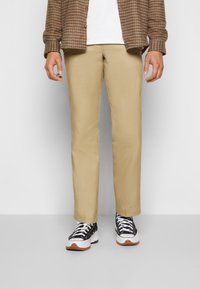 Beige Baumwoll-Chinos mit geradem Bein, kombiniert mit schwarz-weißen Sneakers mit brauner Sohle. Zubehör umfasst ein karierter Hemd.