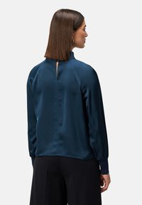 Dunkelblaue Satinbluse mit hohem Kragen und Schlitzöffnung am Rücken. Mit langen Ärmeln und enganliegenden Manschetten sowie einer glatten Textur ausgestattet.