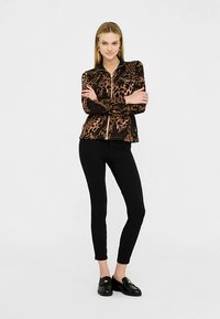 Leopardmuster-Reißverschlussjacke in Braun und Schwarz, kombiniert mit schwarzen Slim-Fit-Hosen und schwarzen Loafers. Glatter Stoff und figurnaher Schnitt.