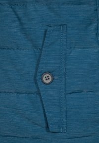 Tissu matelassé bleu avec des coutures horizontales et un rabat cousu maintenu par un seul bouton noir à quatre trous.