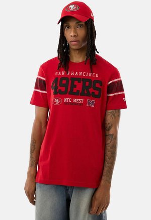 Persona con dreadlocks che indossa un cappellino rosso dei San Francisco 49ers e una maglietta della squadra abbinata con testo e grafica in bianco e nero.
