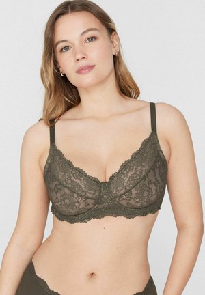 Kobieta o jasnej cerze i blond włosach prezentująca ciemnozielony koronkowy biustonosz z cienkimi ramiączkami na gładkim jasnym tle.