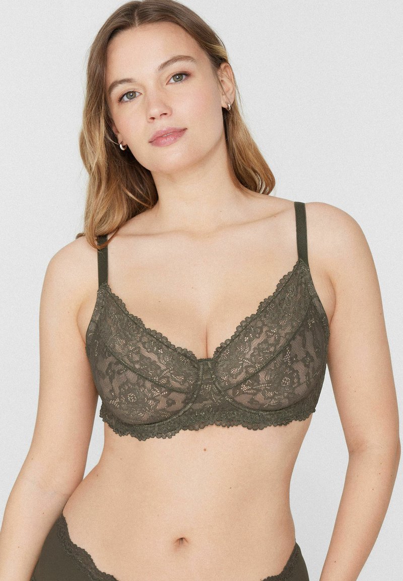 Vrouw met een lichte huid en blond haar die een donkergroene kanten beha met dunne bandjes modelleert tegen een effen lichte achtergrond.