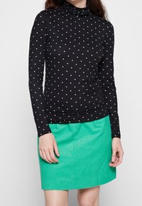 Svart långärmad polotröja med vita prickar, matchad med en grön kordkjol. Toppen har en figurnära design och en slät yta.