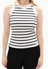 Kvinna iförd en ärmlös, ribbad vit linne med svarta horisontella ränder och svarta byxor mot en vit bakgrund.