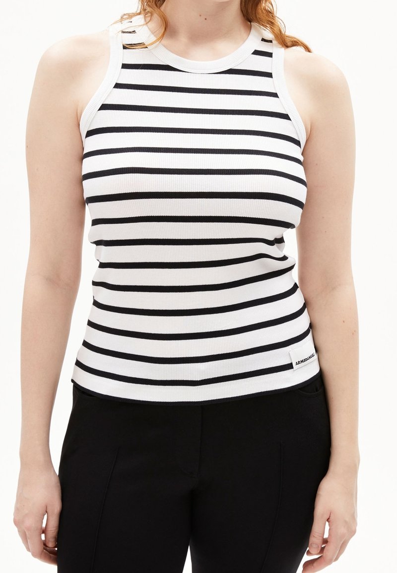 Kvinna iförd en ärmlös, ribbad vit linne med svarta horisontella ränder och svarta byxor mot en vit bakgrund.