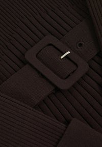 Bruin ribbelstof met een brede riem met een vierkante gesp en een witte spelddetail, wat een gestructureerd, textuurachtig uiterlijk creëert.