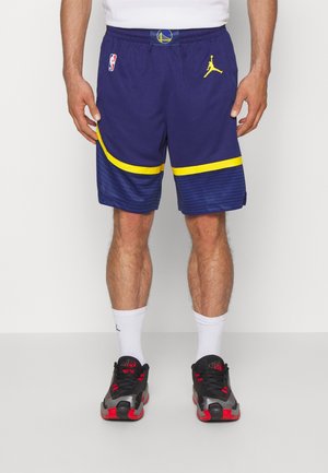 Man draagt blauwe en gele Golden State Warriors basketbalshorts, witte sokken en zwart-rode sportschoenen, staand met de handen langs het lichaam.
