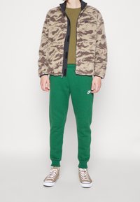 Giacca in pile beige con motivo mimetico marrone, abbinata a pantaloni della tuta verdi e sneakers color sabbia con suole bianche. Chiusura frontale con cerniera.