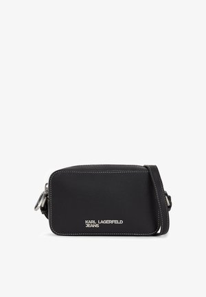 Juoda sintetinio odos crossbody rankinė su balta siūle ir sidabriniais aksesuarais. Pagrindinis bruožas - prekės ženklo logo priekyje ir reguliuojama dirželis.