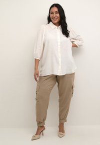Chemise blanche à boutons avec col et manches bouffantes, associée à un pantalon cargo beige avec poches latérales et poignets élastiques.
