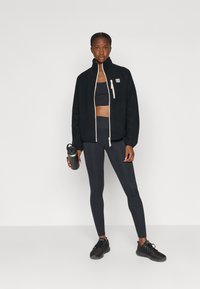 Jachetă din fleece neagră cu fermoar, cu accente albe, sutien sport negru, colanți închiși la culoare și adidași negri; ținând o sticlă de apă gri.