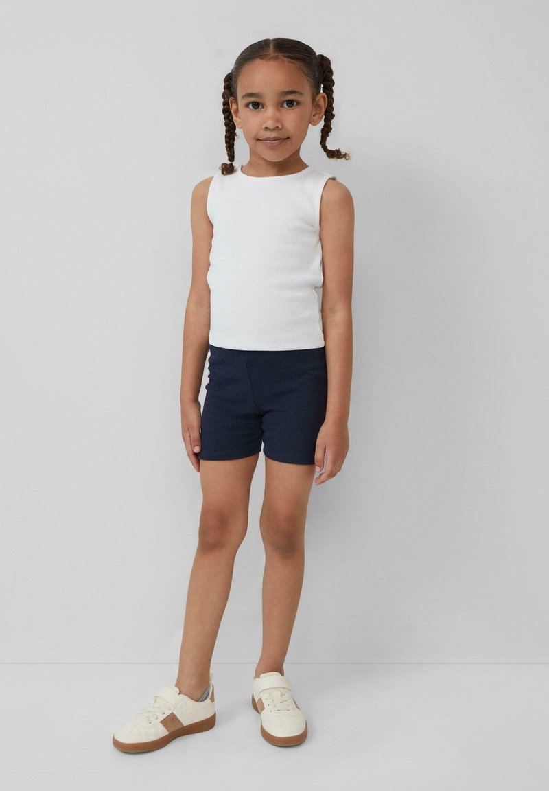 Jong meisje met gevlochten haar, gekleed in een witte mouwloze top, marineblauwe shorts en witte sneakers, staand tegen een effen grijze achtergrond.