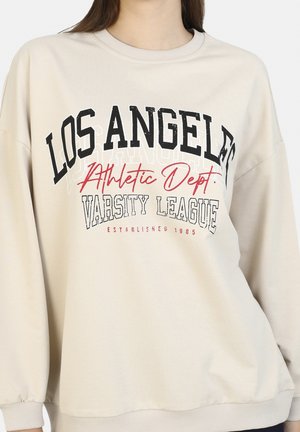 Sweat-shirt beige avec le texte "LOS ANGELES Athletic Dept. VARSITY LEAGUE Established 1985" en lettres noires, rouges et blanches sur la poitrine.