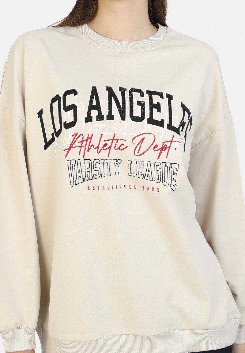 Sweat-shirt beige avec le texte "LOS ANGELES Athletic Dept. VARSITY LEAGUE Established 1985" en lettres noires, rouges et blanches sur la poitrine.
