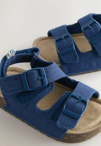 Gros plan sur des sandales bleues pour tout-petit avec des boucles ajustables et des semelles en liège sur un fond clair.