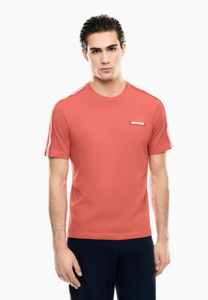 Mand iført en koralfarvet T-shirt med korte ærmer med sidelommer og mørke bukser, stående mod en hvid baggrund.
