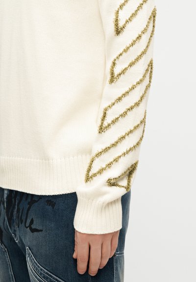 Μανίκι από πουλόβερ πλεκτό σε κρεμ χρώμα με υφασμένες ραβδώσεις σε σχήμα ζιγκ-ζαγκ σε ελιά πράσινο, φορεμένο με σκούρο μπλε τζιν τζιν παντελόνι που δείχνει μαύρο τυπωμένο κείμενο.