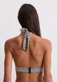 Mujer con cabello largo y castaño, vistiendo un top de bikini de flores en blanco y negro, vista desde atrás contra un fondo blanco sencillo.
