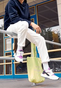 Weiße Sneaker mit lila Akzenten, Überlagerungsdesign, schwarze Sohle. Das Modell trägt weiße Hosen, eine grüne Tragetasche mit Text und einen marineblauen Hoodie.